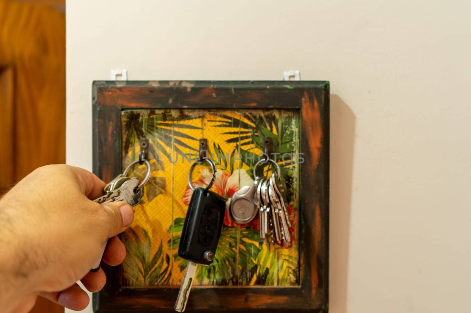 Man taking door key from wooden key holder hanging on the white wall, close up shot of male's hand.