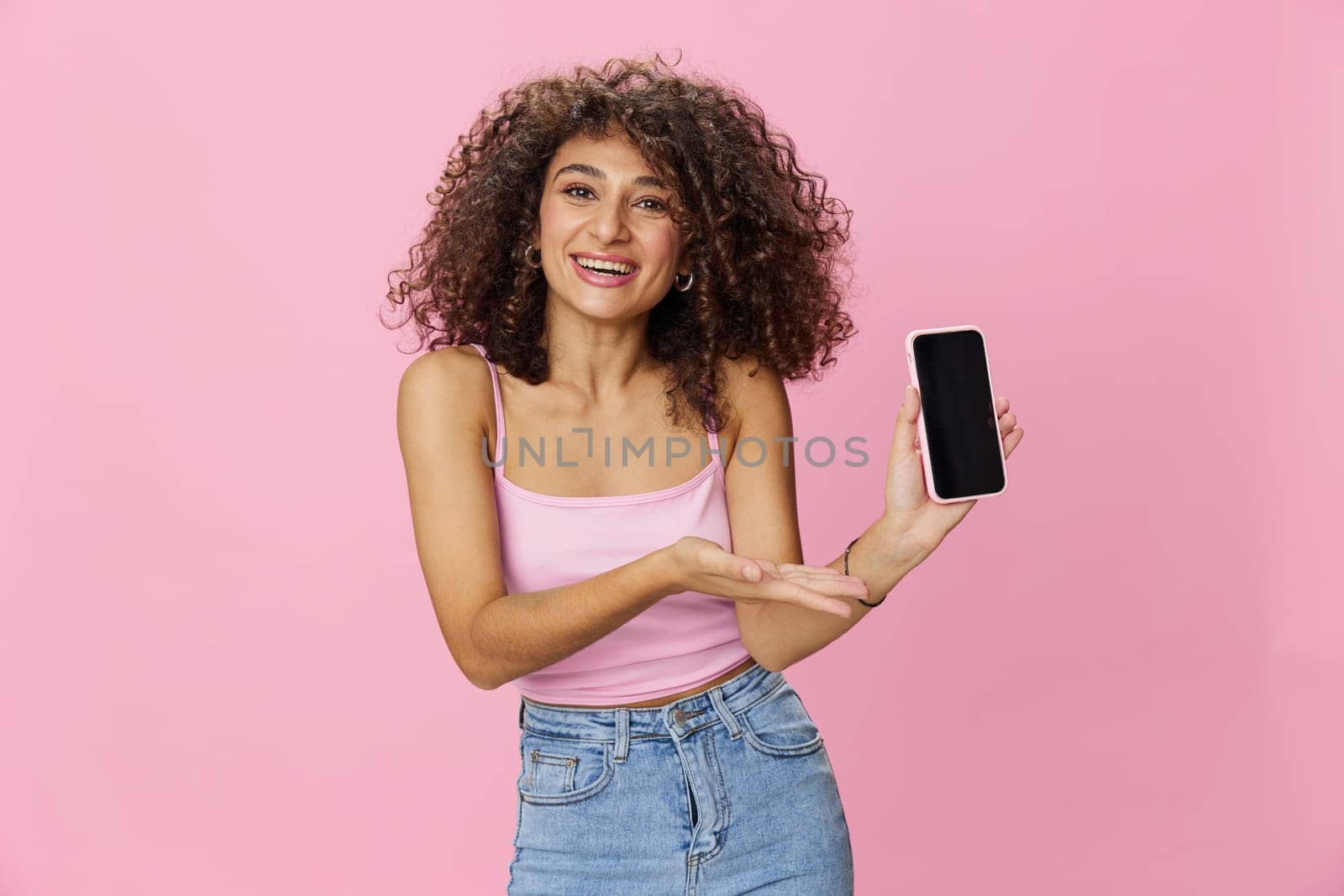 Woman blogger holding phone video call takes selfies, with curly hair in pink smile t-shirt and jeans poses on pink background, copy space, technology and social media, online. High quality photo