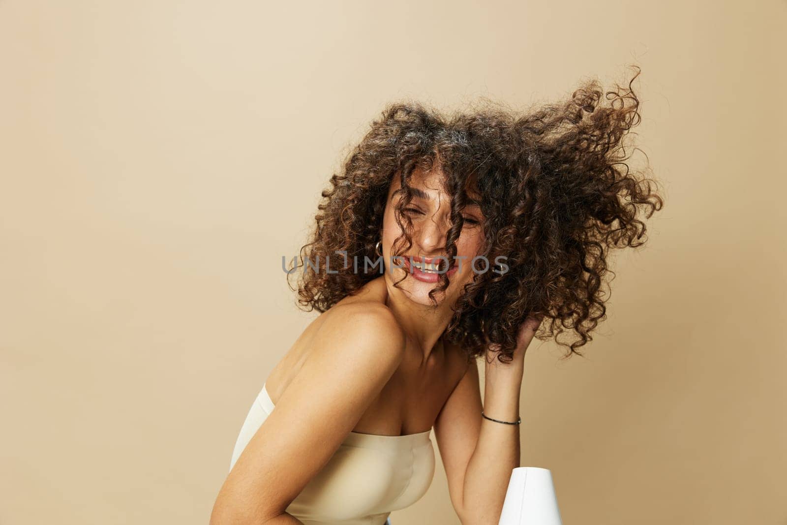 Woman dries curly afro hair with blow dryer, home beauty care styling products hair, smile on beige background by SHOTPRIME