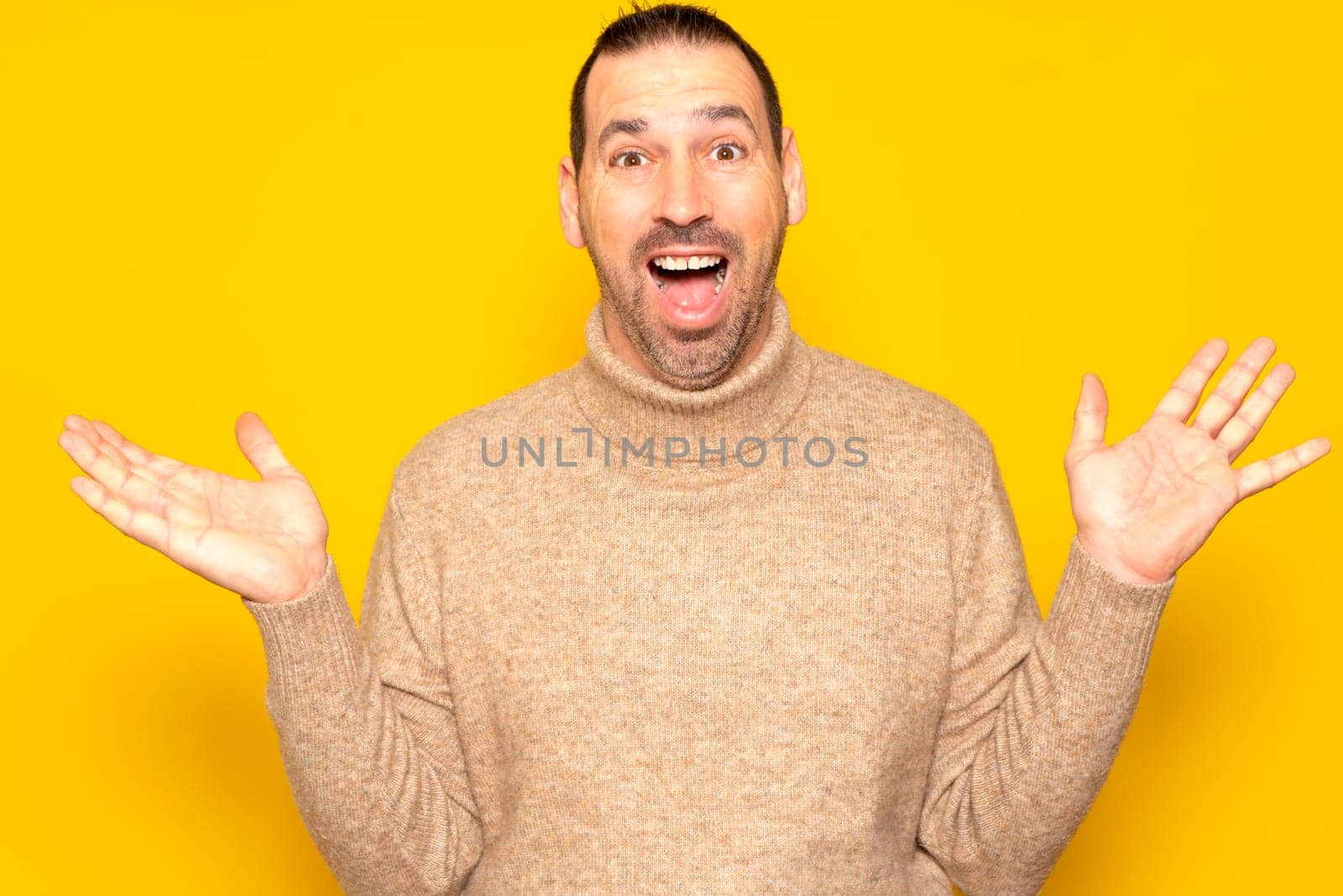 Funny smiling man in beige sweater showing palms up, unexpected surprise gesture isolated over yellow background. Attractive happy man listening to good news, catching something