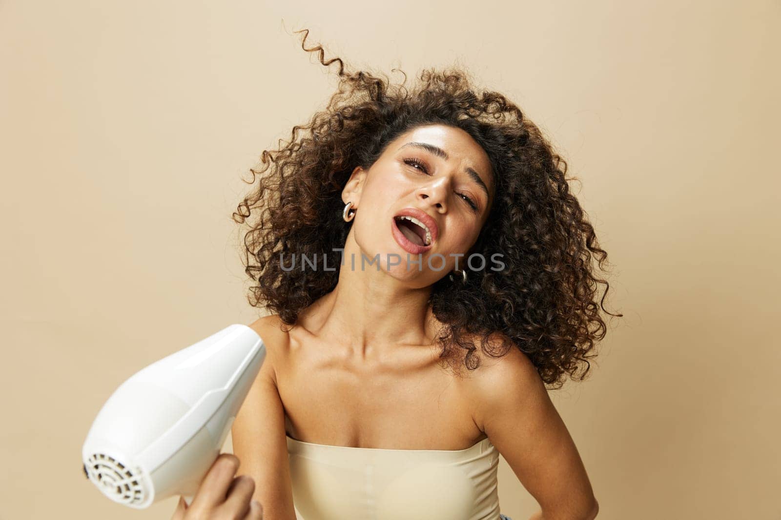 Woman dries curly afro hair with blow dryer, home beauty care styling products hair, smile on beige background by SHOTPRIME