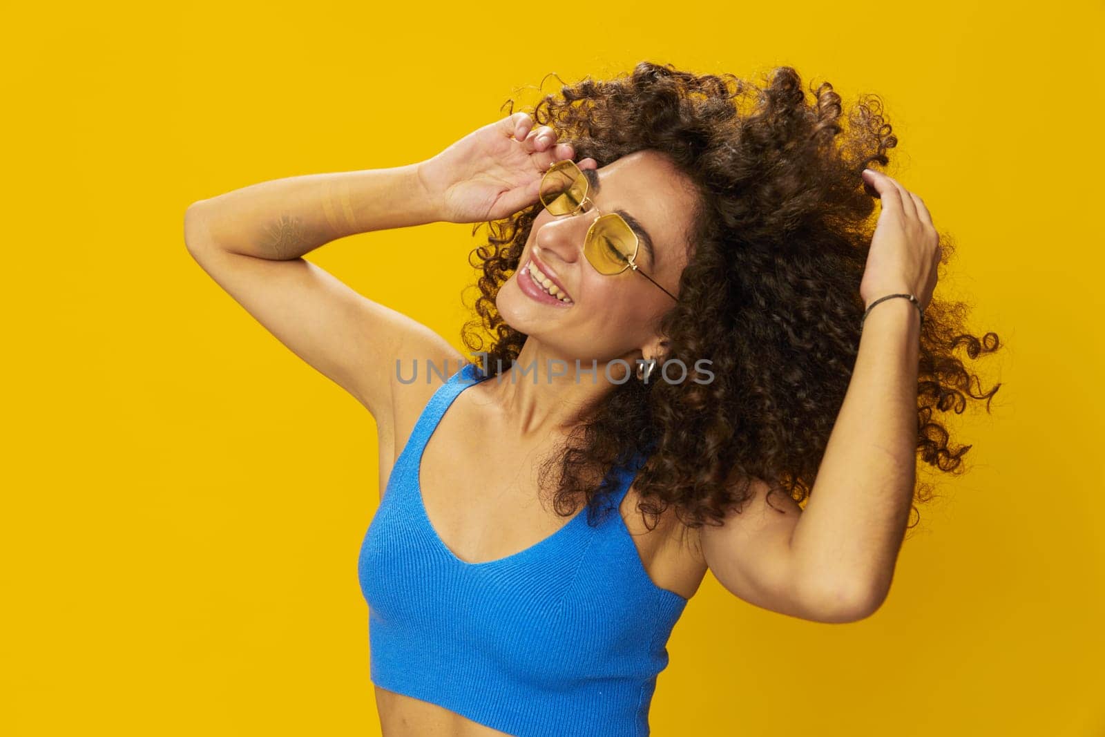 Woman with curly afro hair in a blue T-shirt on a yellow background dancing flying hair with sunglasses yellow, hand signs, look into the camera, smile with teeth and happiness, copy space by SHOTPRIME