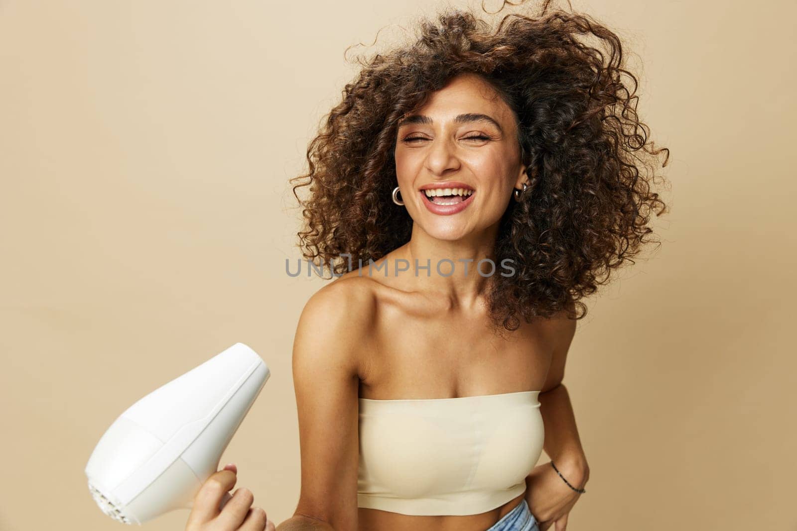 Woman dries curly hair with blow dryer, home beats styling products, smile on beige background. High quality photo