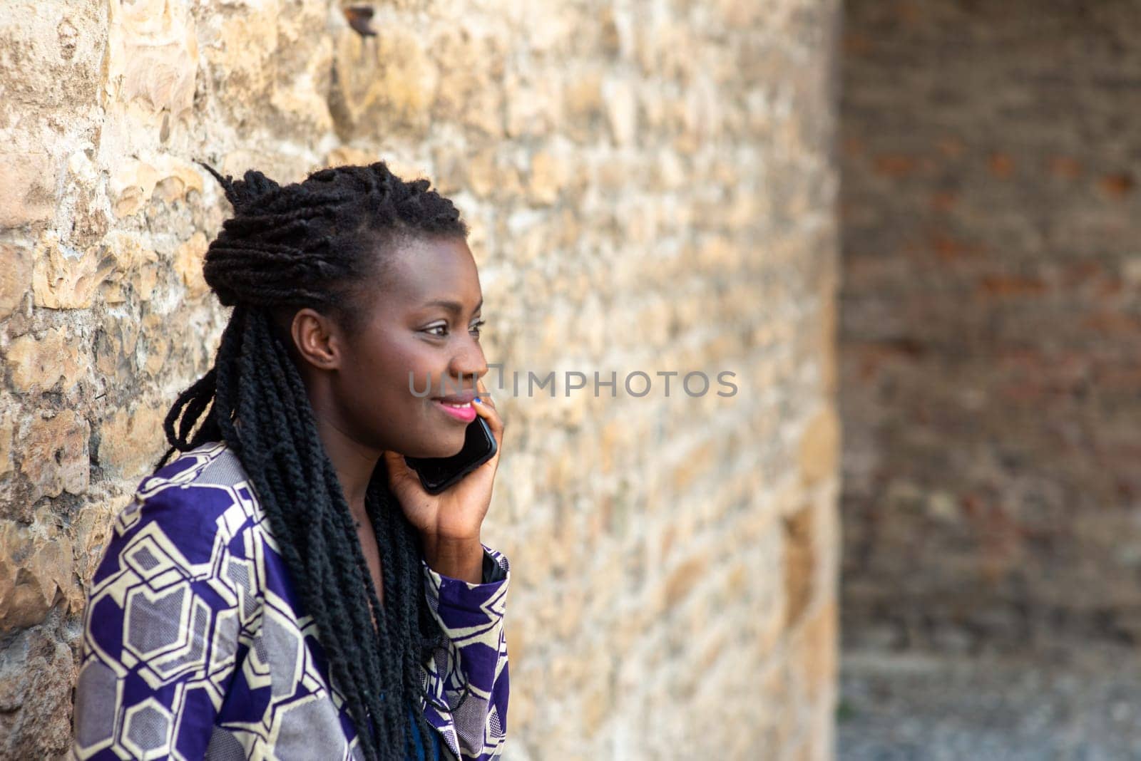 confident African young adult woman holding cell phone calling , receives good news, smiling, happy, outdoors in the village over a wall.