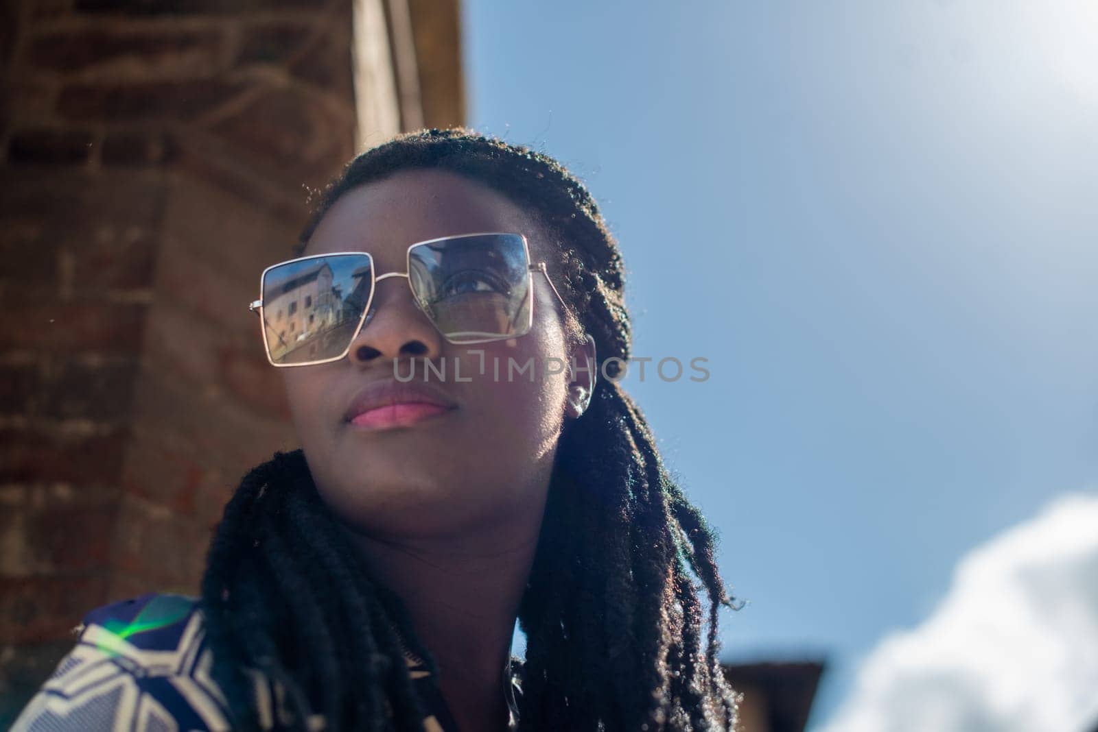 Happy young woman feeling confident in her style. Fashionable woman wearing sunglasses and braided hairstyle outdoors. Tourist traveling italian old village.