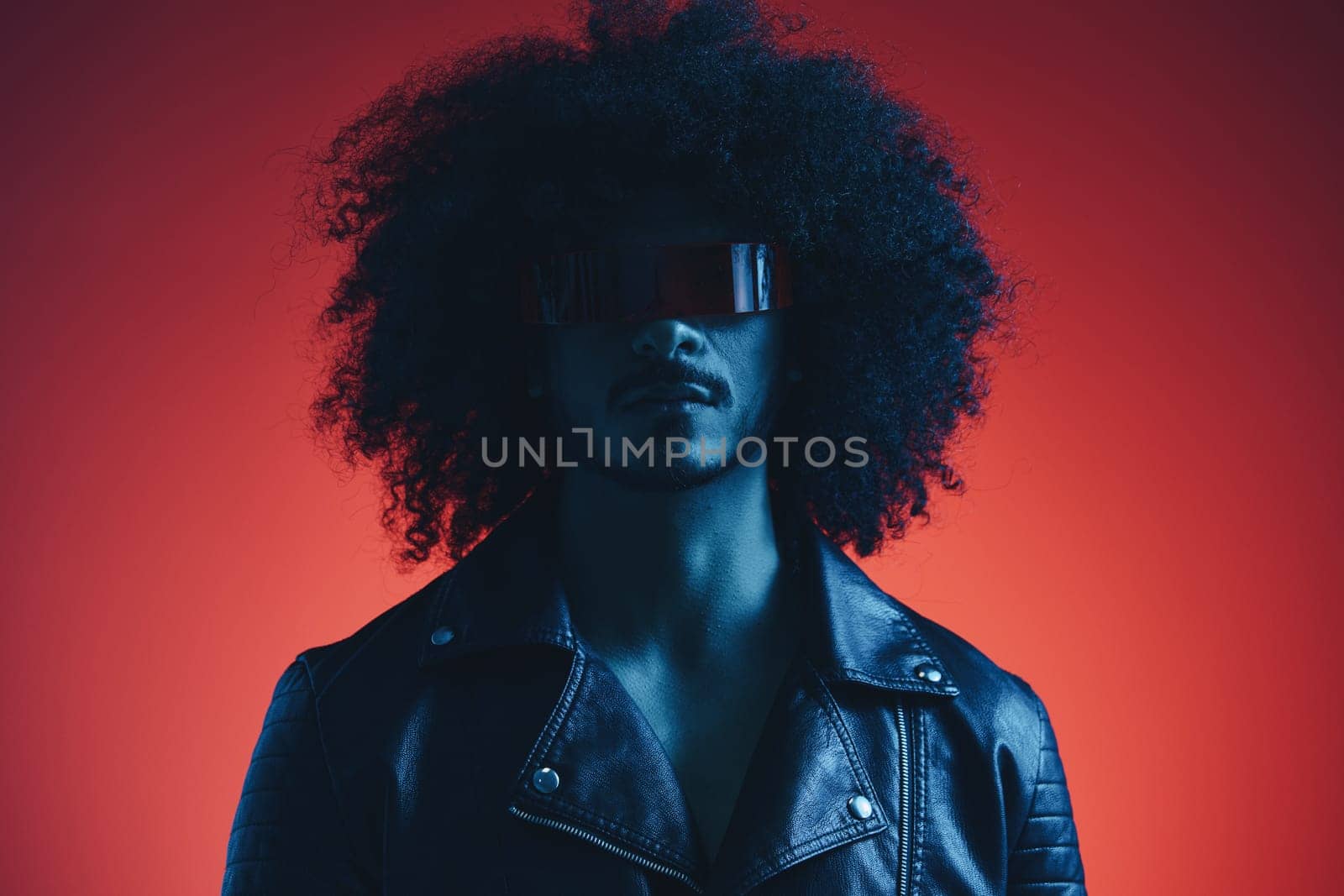 Portrait of fashion man with curly hair on red background with stylish glasses, multicultural, colored light, black leather jacket trend, modern concept. High quality photo
