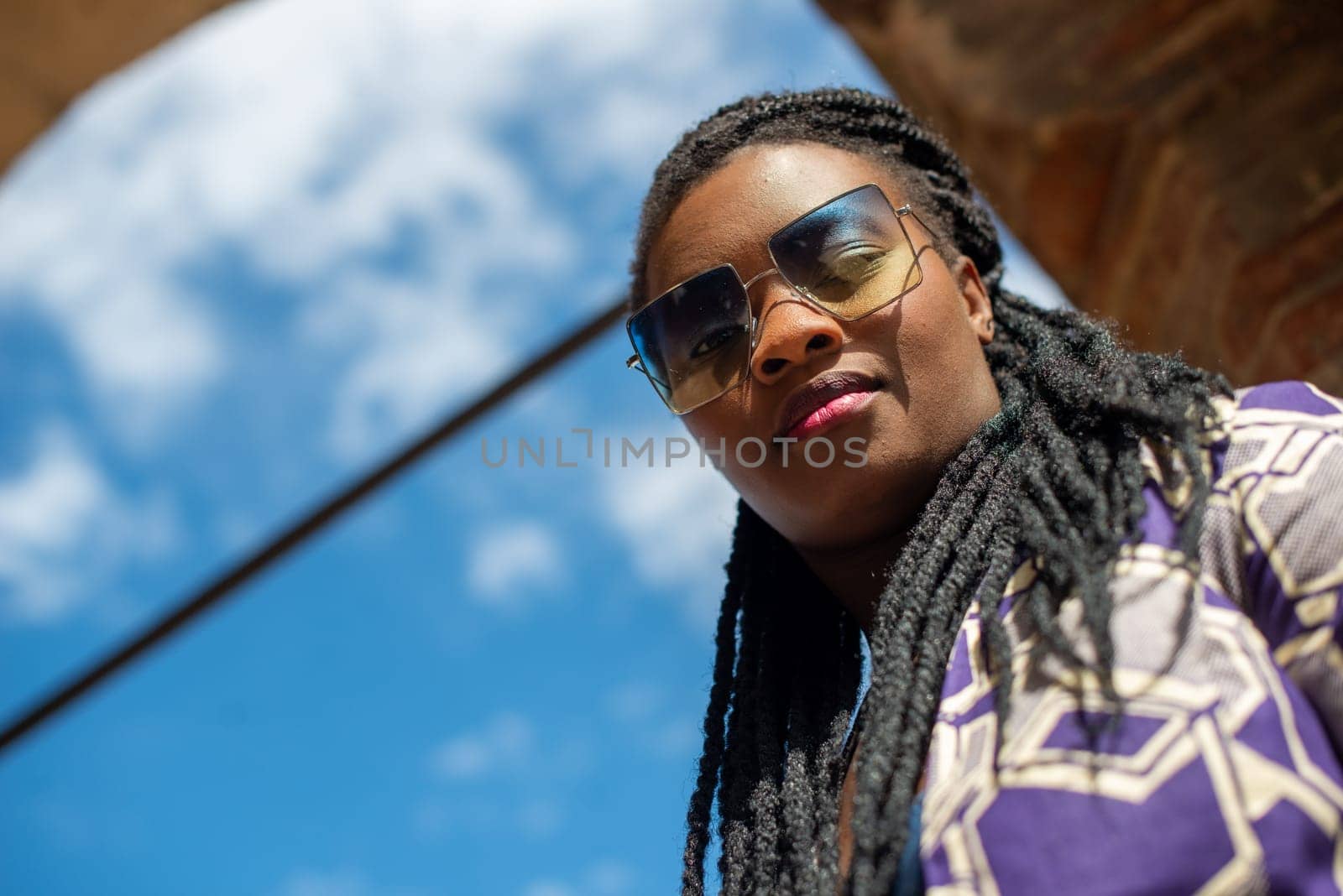 Happy young woman feeling confident in her style. Fashionable woman wearing sunglasses and braided hairstyle outdoors. Tourist traveling italian old village.