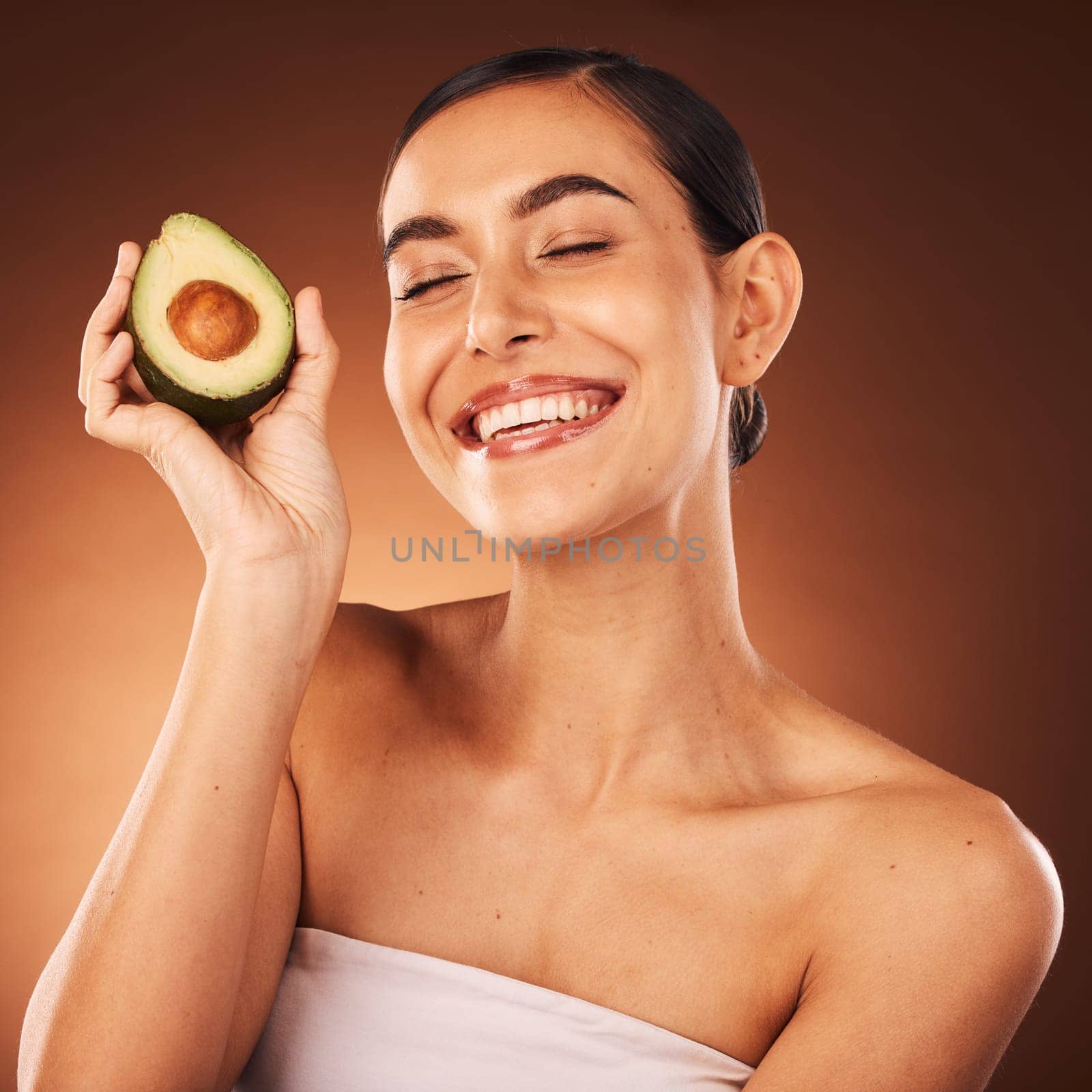 Face, skincare and avocado with a model woman in studio on a brown background to promote healthy eating. Food, health and wellness with an attractive young female inside for luxury or natural care by YuriArcurs