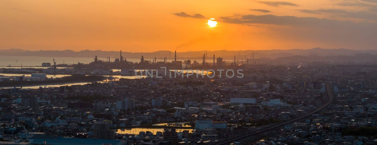 Panoramic aerial view of low sun in orange sky over industrial smokestacks and city at sunset. High quality photo