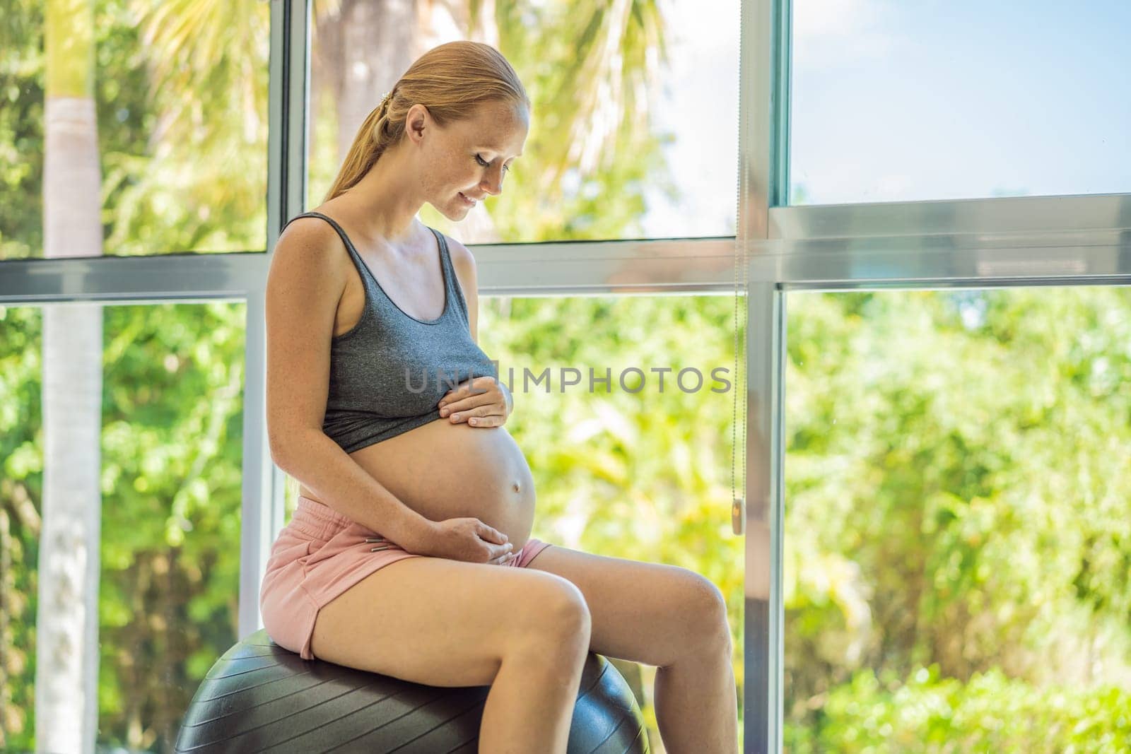 Pregnant woman exercising on fitball at home. Pregnant woman doing relax exercises with a fitness pilates ball. Against the background of the window.