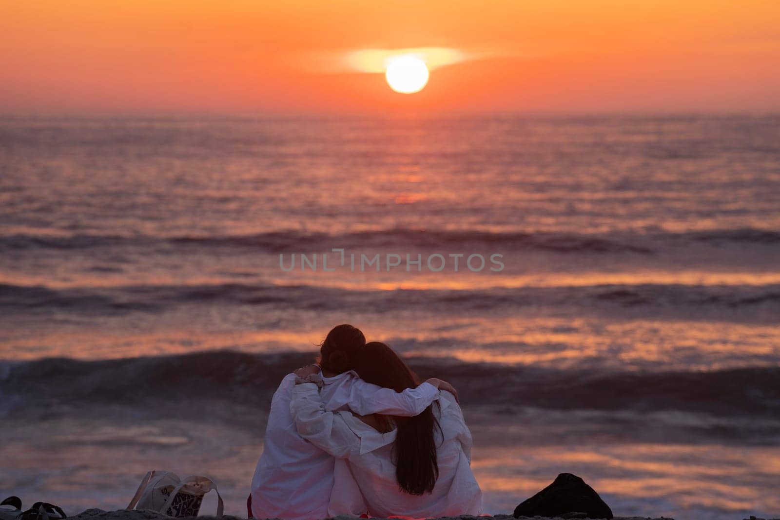 Rear view of a couple silhouette sitting cuddling and enjoying pointing at sun at sunset outside on the beach by papatonic