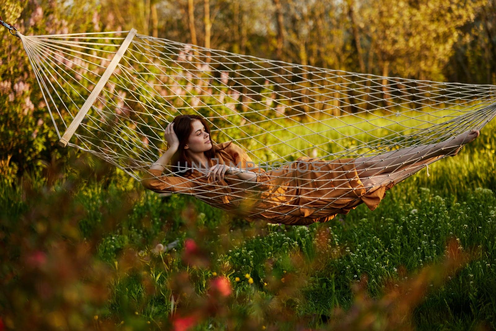 a beautiful woman is resting in nature lying in a mesh hammock in a long orange dress looking to the side, propping her head with her hand. Horizontal photo on the theme of recreation. High quality photo