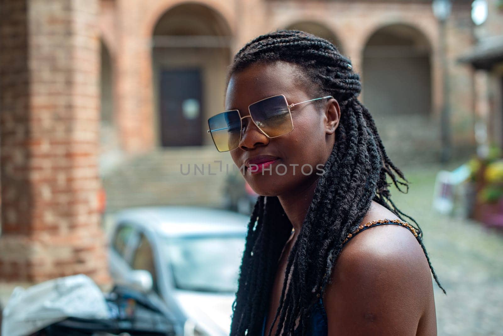 Happy young woman feeling confident in her style. Fashionable woman wearing sunglasses and braided hairstyle outdoors. Tourist traveling italian old village.
