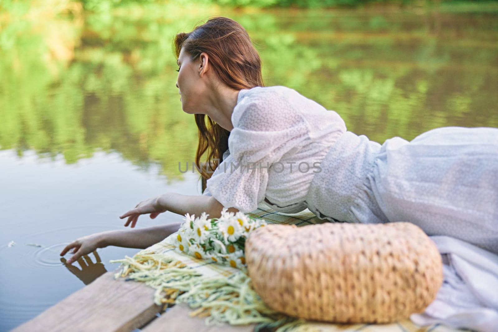 a woman in a light dress lying on a pier by the lake touches the water by Vichizh