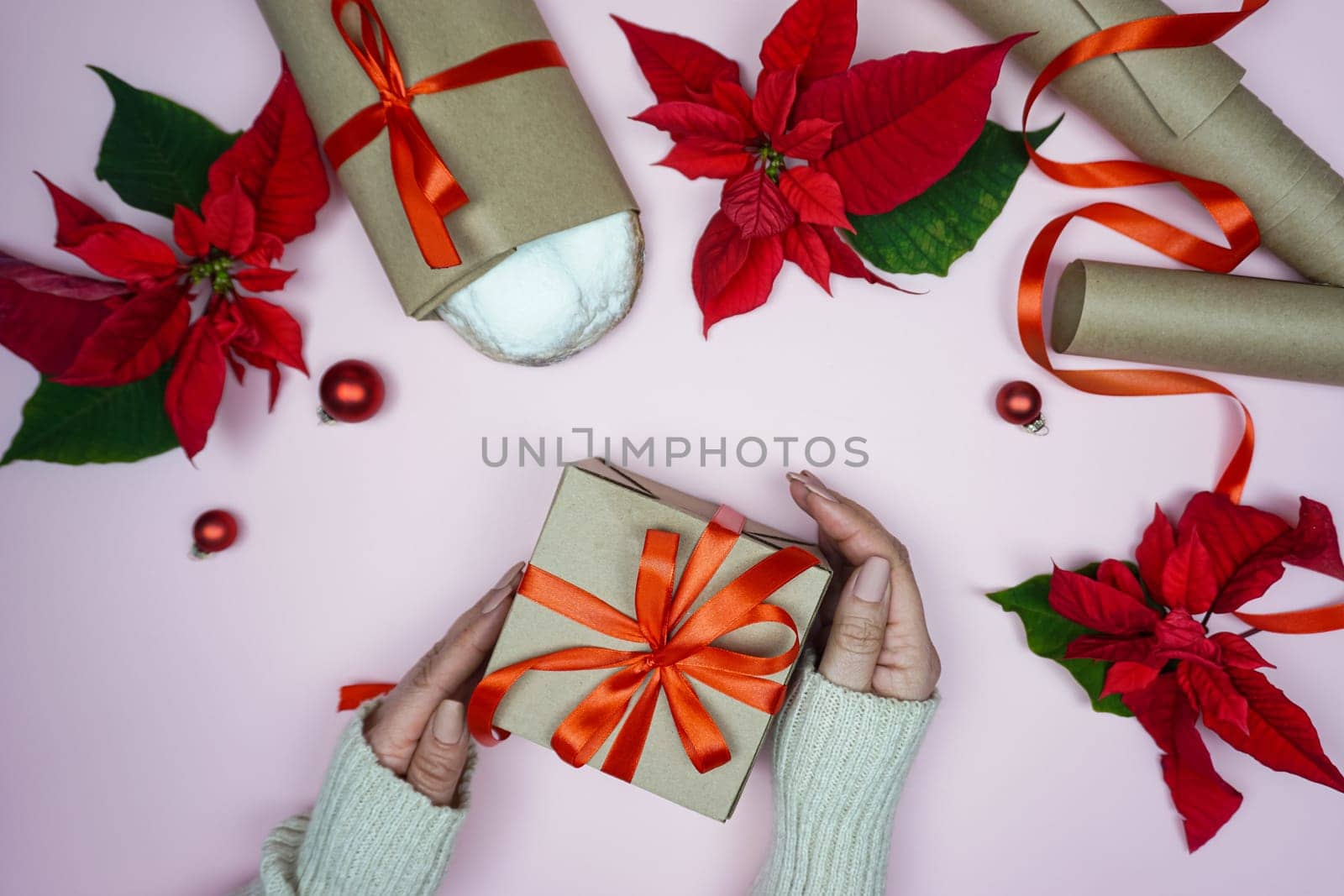 In women's hands, a gift in craft paper and with a red bow. On a pink background, Christmas items are laid out - stollen, Poinsettia flower, craft paper for wrapping gifts.