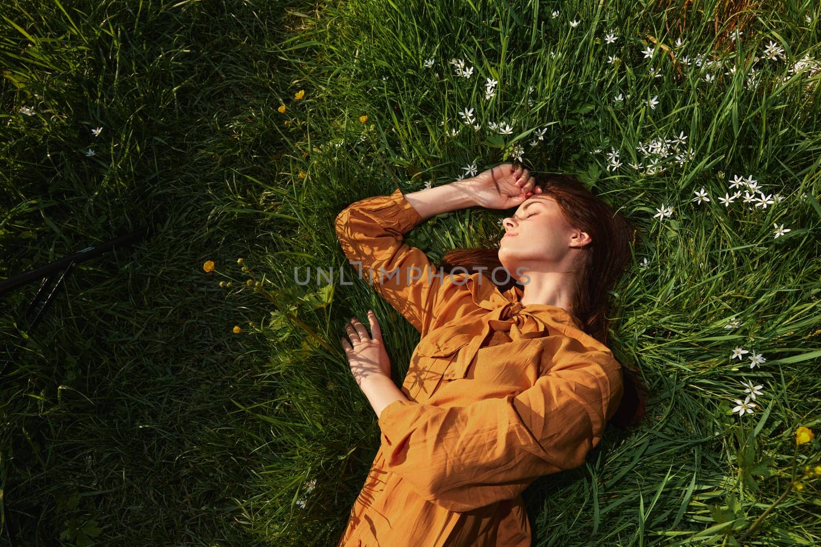 horizontal photo of a happy, relaxed woman, resting lying in the grass, in a long orange dress, with closed eyes and a pleasant smile on her face, enjoying harmony with nature and recuperating by Vichizh