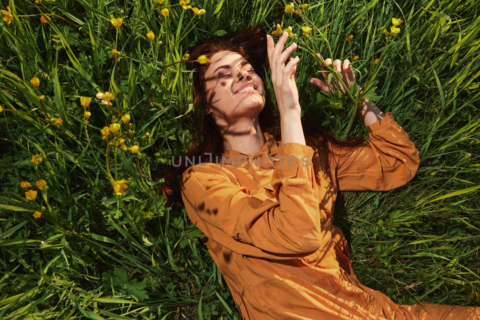 a calm woman with long red hair lies in a green field with yellow flowers, in an orange dress with her eyes closed, touching blades of grass with her hands, enjoying peace and recuperating by Vichizh