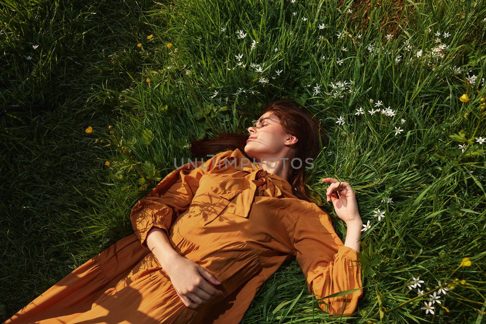 a calm woman with long red hair lies in a green field with yellow flowers, in an orange dress with her eyes closed, with a pleasant smile on her face, enjoying peace, illuminated by the setting sun by Vichizh