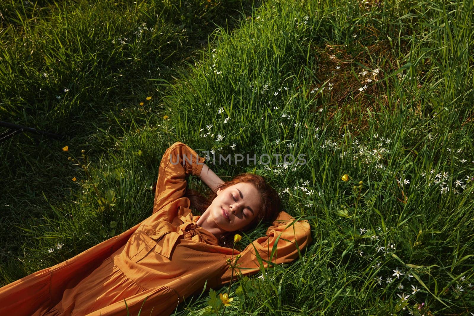 a happy, relaxed woman, resting lying in the green grass, in a long orange dress, with her eyes closed and a pleasant smile on her face, enjoying harmony with nature and recuperating by Vichizh