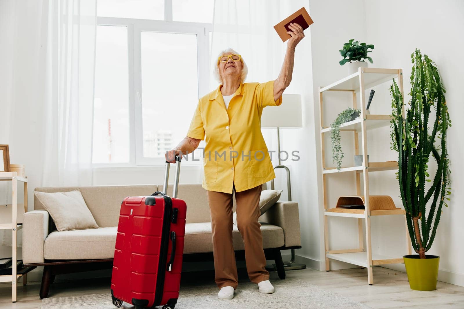 Happy senior woman with passport and travel ticket packed a red suitcase, vacation and health care. Smiling old woman joyfully stands in the house before the trip. by SHOTPRIME
