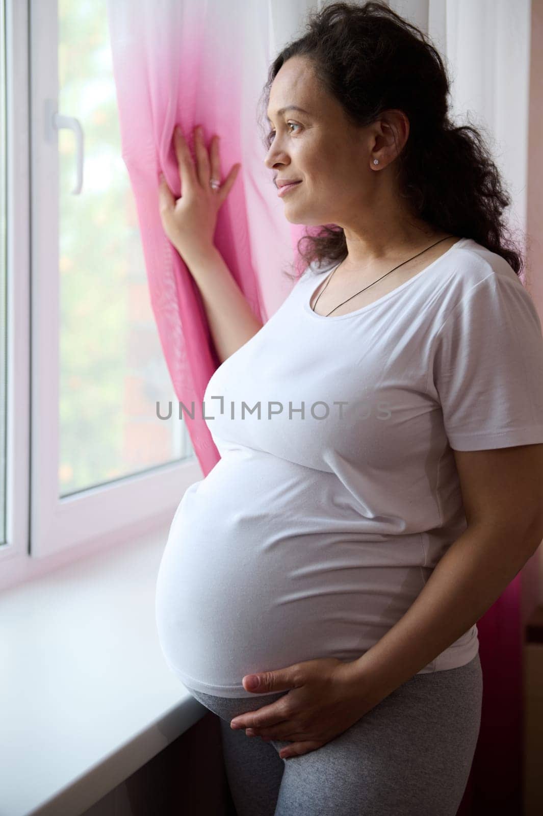 Beautiful curly multi-ethnic pregnant woman, expectant mother in white t-shirt, dreamily looking out the window, touching her big belly in late pregnancy, feeling harmony, enjoying maternity lifestyle