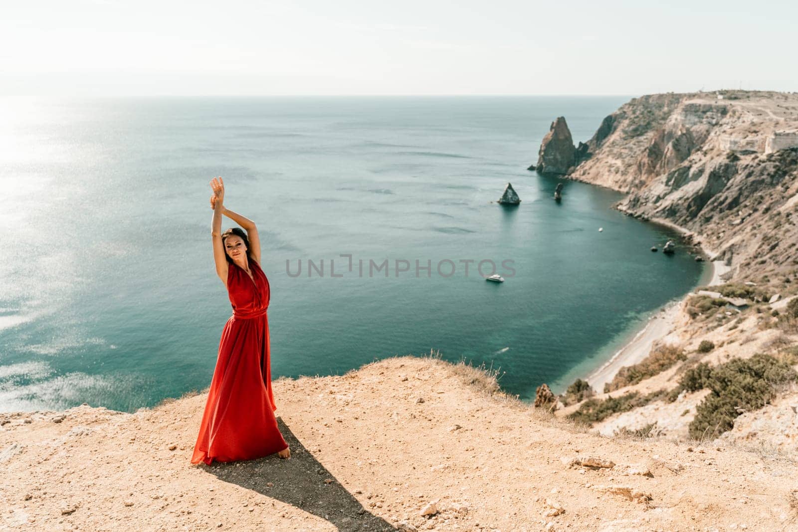 Woman red dress sea. posing on a rocky outcrop high above the sea. Girl on the nature on blue sky background. Fashion photo. by Matiunina