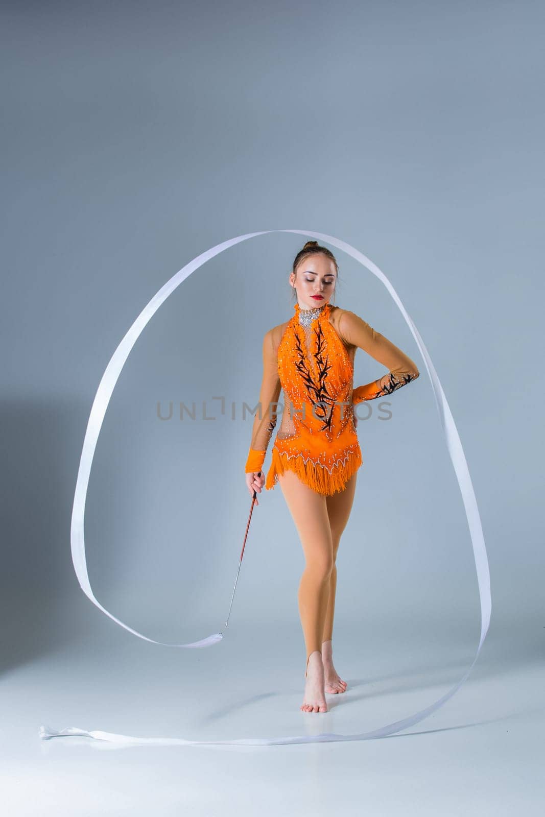 teenager doing gymnastics dance with white ribbon on a blue background. The girl in the orange dancing suit