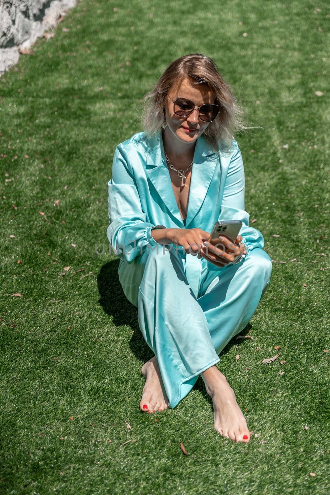 Woman grass. Happy woman with blond curly hair in glasses and in a blue overalls sits on the summer fresh green grass with a phone in her hands