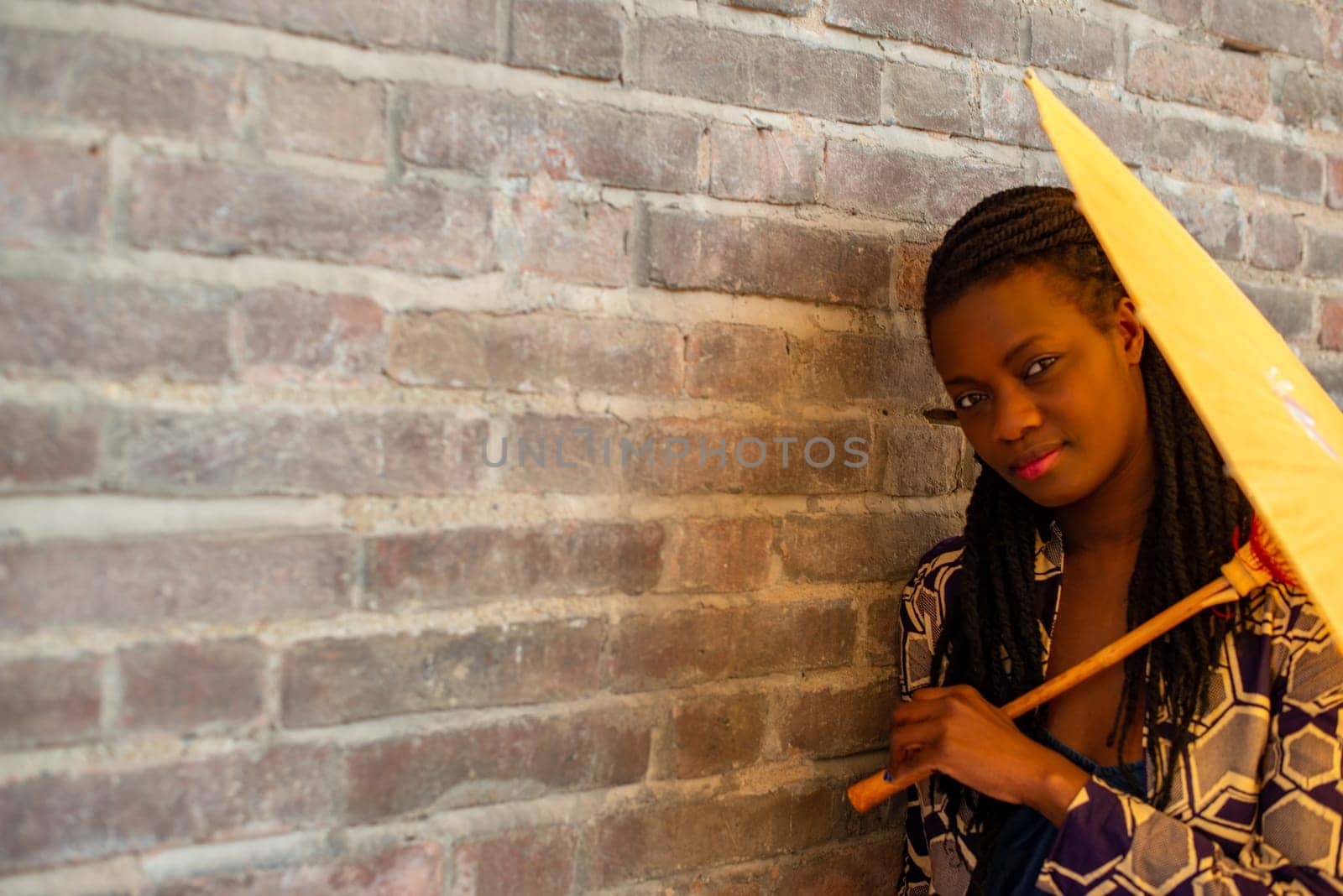 Young woman with dreadlocks braided hairstyle holding umbrella parasol. Happy young woman feeling confident in her style. Fashionable woman standing in the street of old village against stone wall.