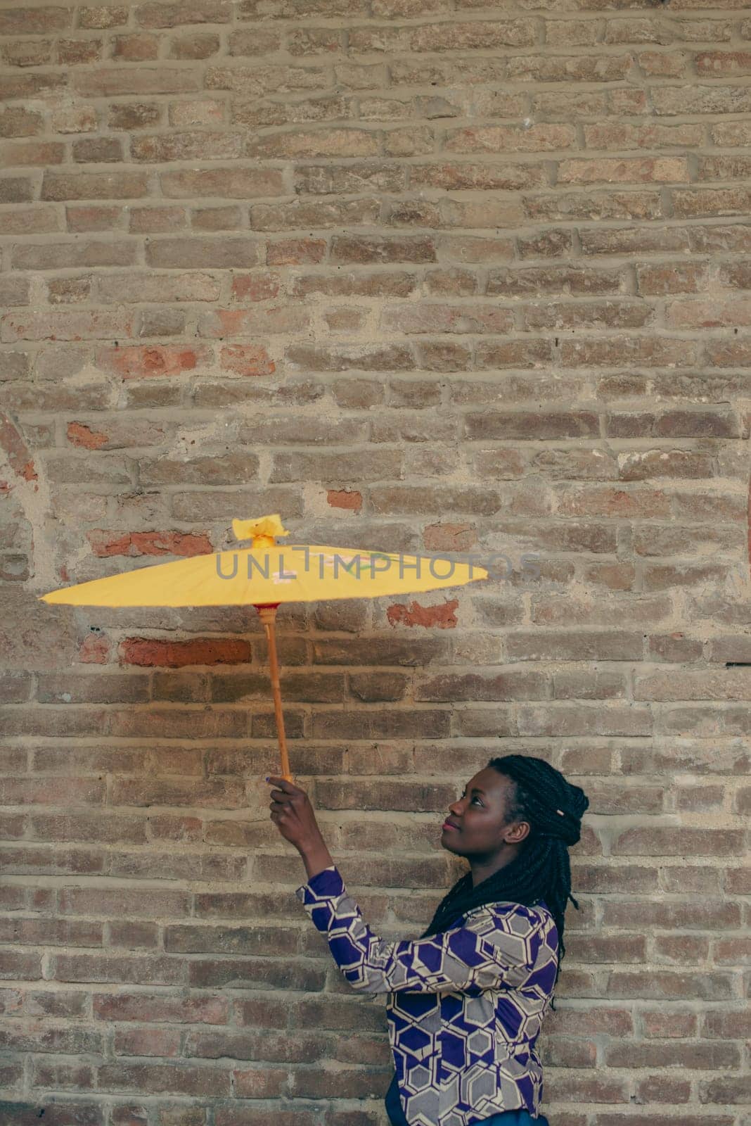 Young woman with dreadlocks braided hairstyle holding umbrella parasol. Happy young woman feeling confident in her style. Fashionable woman standing in the street of old village against stone wall.