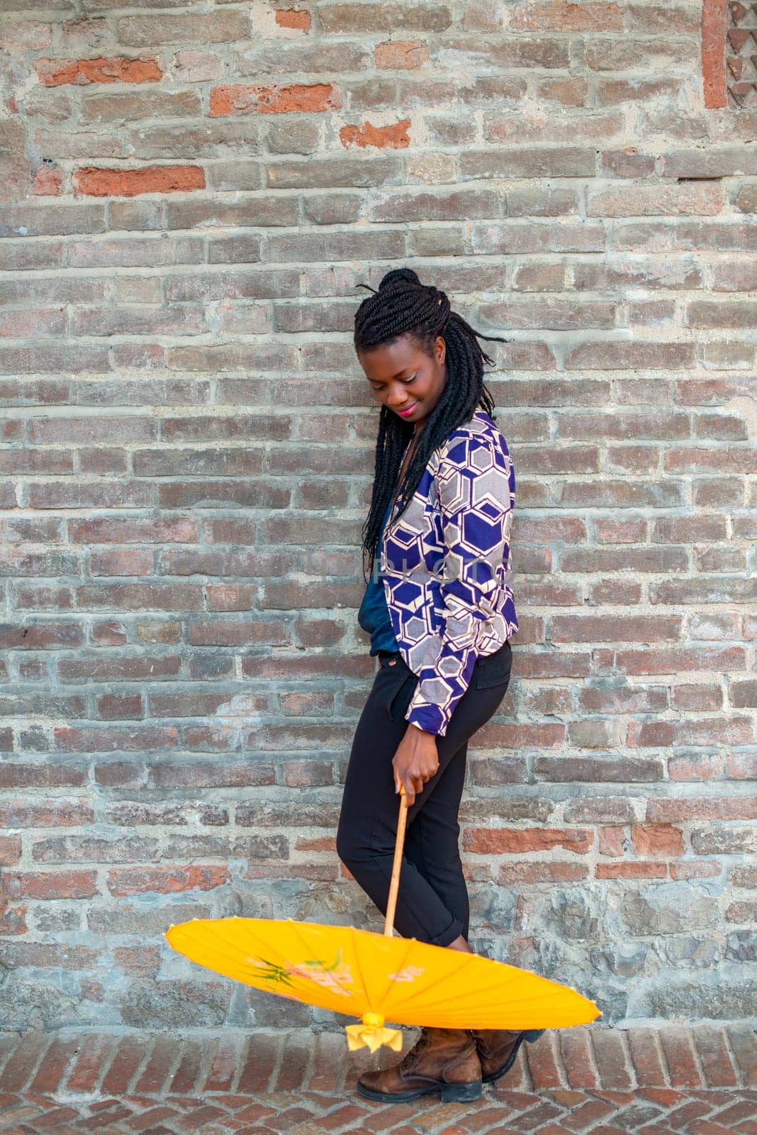 Young woman with dreadlocks braided hairstyle holding umbrella parasol. Happy young woman feeling confident in her style. Fashionable woman standing in the street of old village against stone wall.