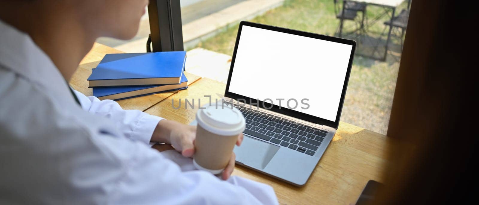 View over shoulder of doctor holding paper cup of coffee and using laptop on wooden table in medical office.