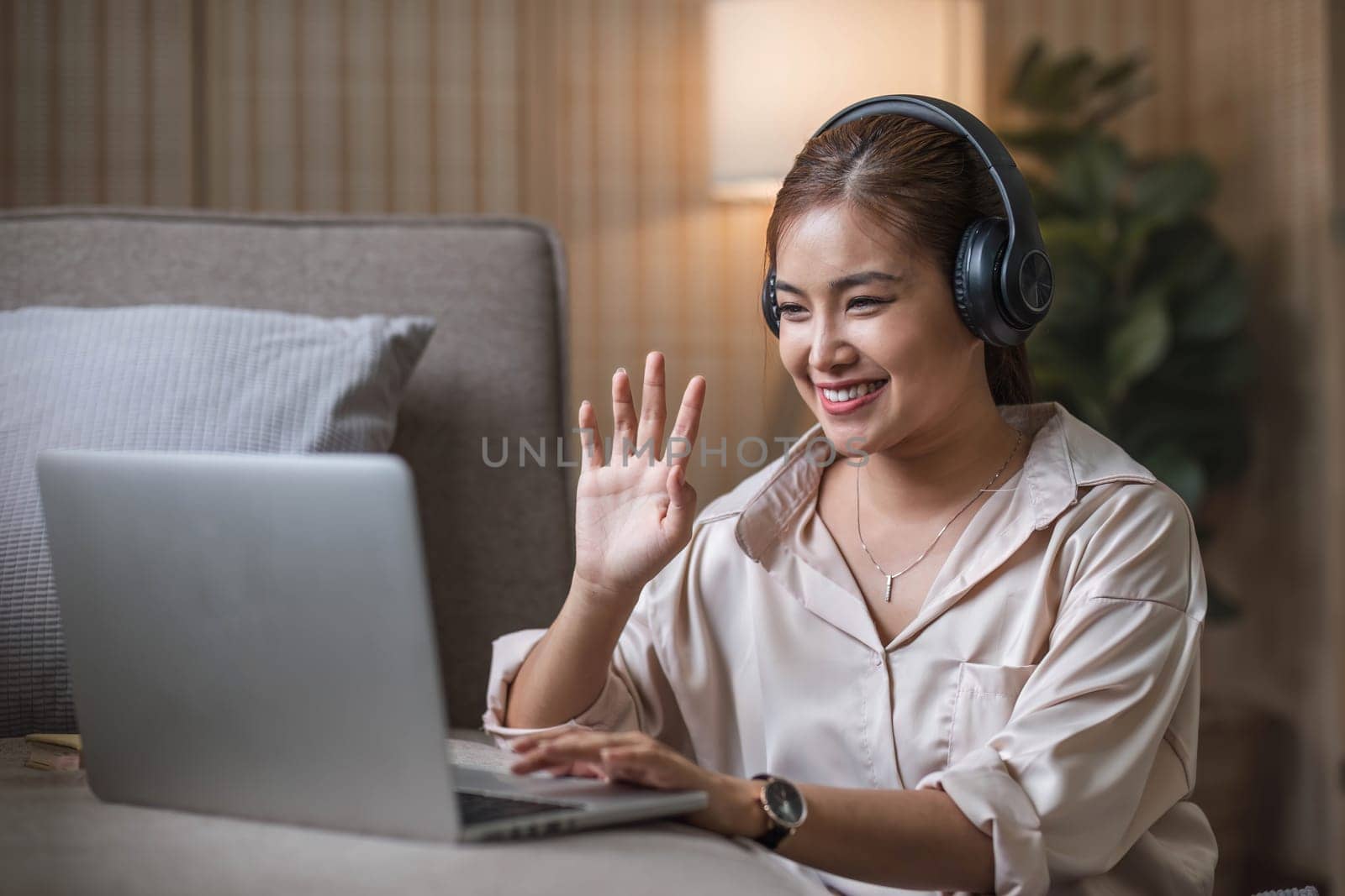 Smiling young asian woman using laptop web camera while wearing headphones and sitting on the rug beside to the sofa at homes..