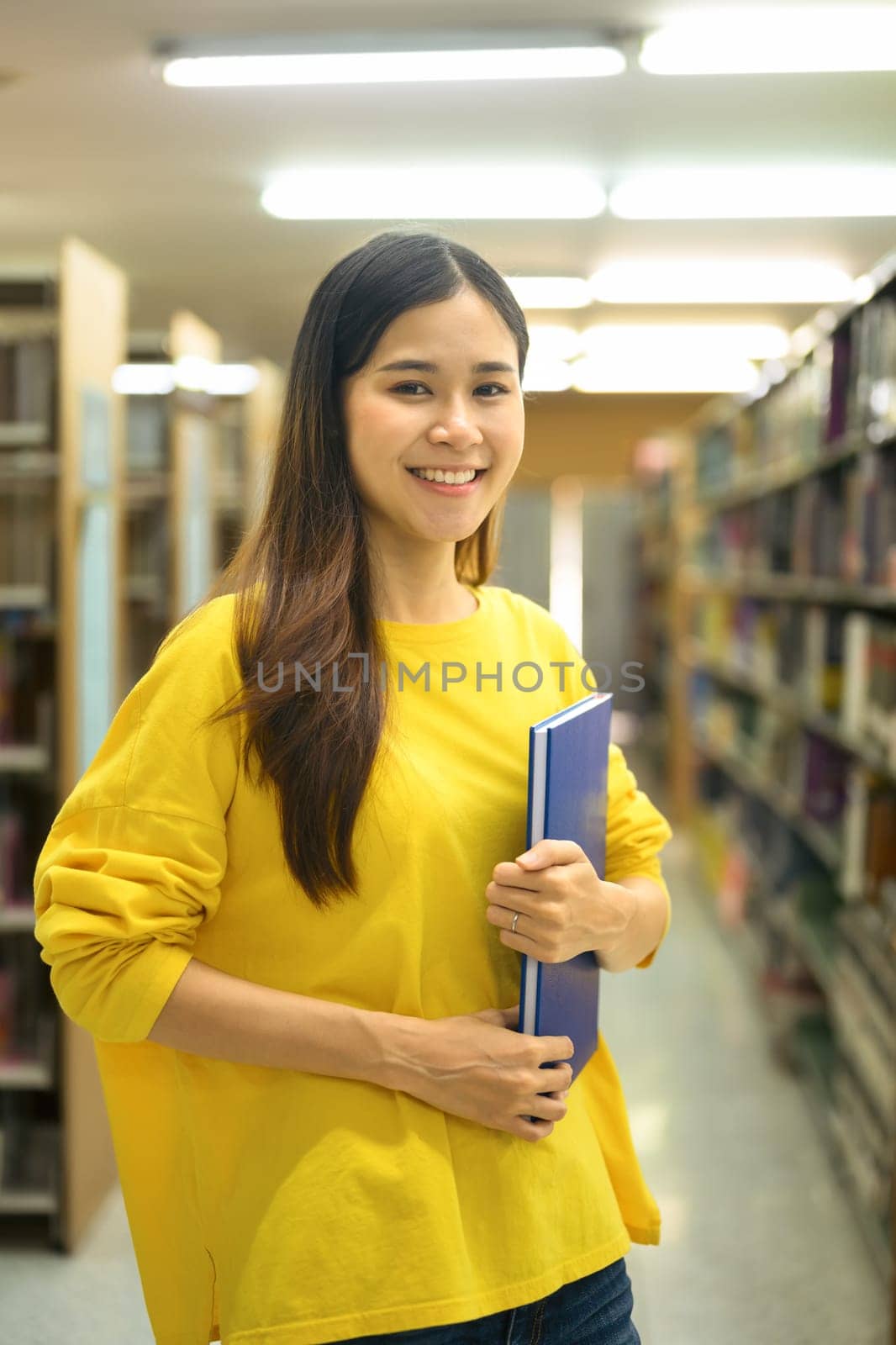 Portrait of university student woman holding textbook standing in library, looking at camera. Knowledge and education concept.