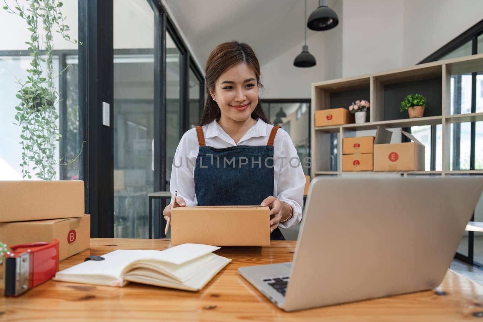 Happy young asian woman startup small business freelance holding parcel box and computer laptop and sitting on chair, Online marketing packing box delivery concept by nateemee