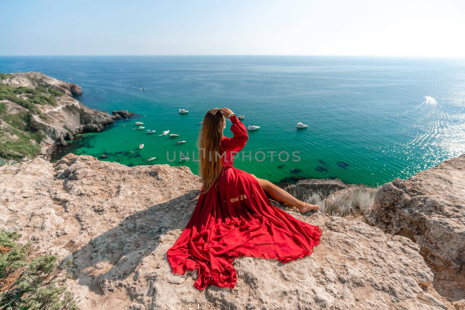 Woman red dress sea. Happy woman in a red dress and white bikini sitting on a rocky outcrop, gazing out at the sea with boats and yachts in the background. by Matiunina