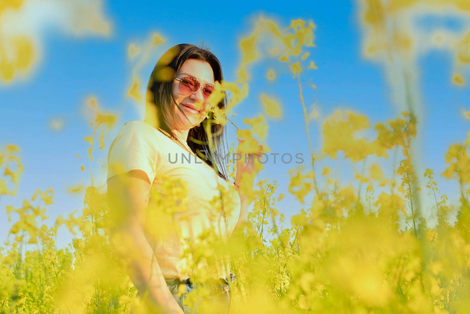 A beautiful girl in sunglasses stands in a field of yellow flowers against a blue sky, smiles happily and inhales the fresh spring and clean air. Peace concept. The color of the flag of Ukraine. No war.