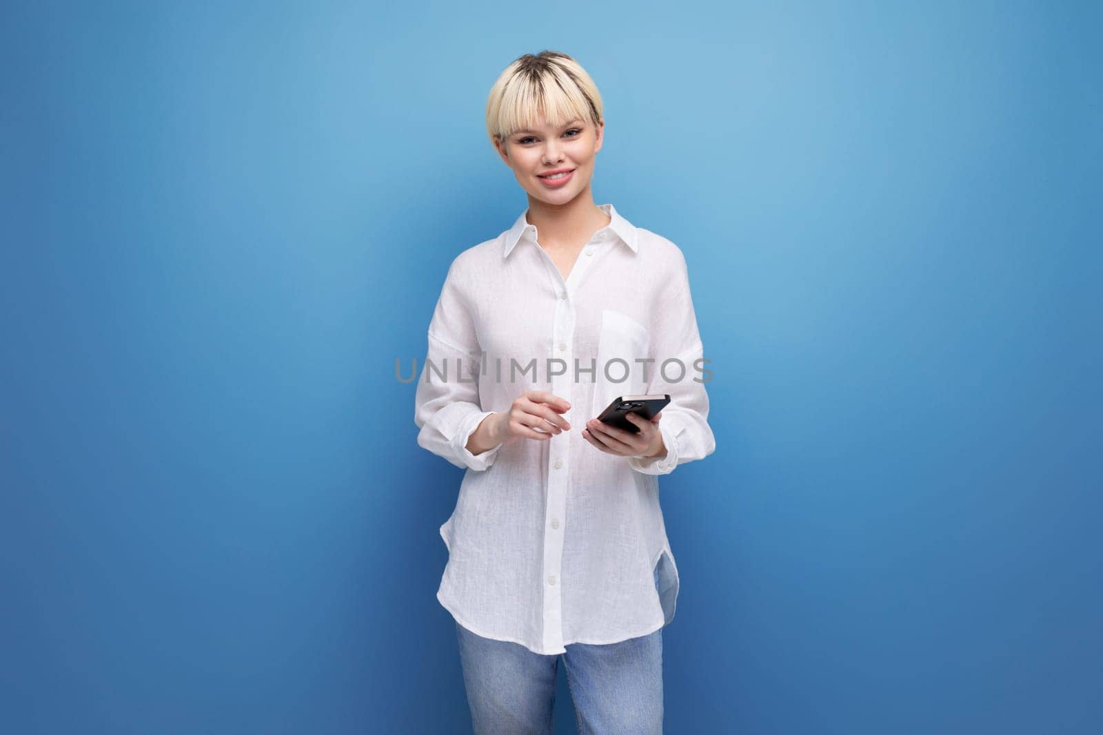 young cheerful pretty blonde businesswoman in a white shirt holds a phone in her hand against the background with copy space.