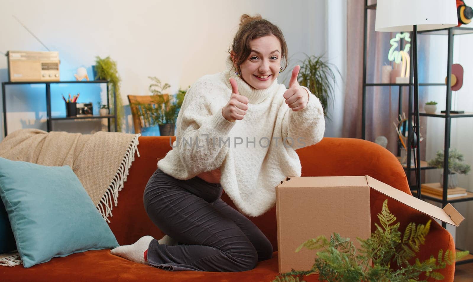 Happy young woman unpacking delivery parcel sitting at home apartment on sofa. Smiling satisfied girl shopper online shop customer opening cardboard box receiving purchase gift by fast postal shipping