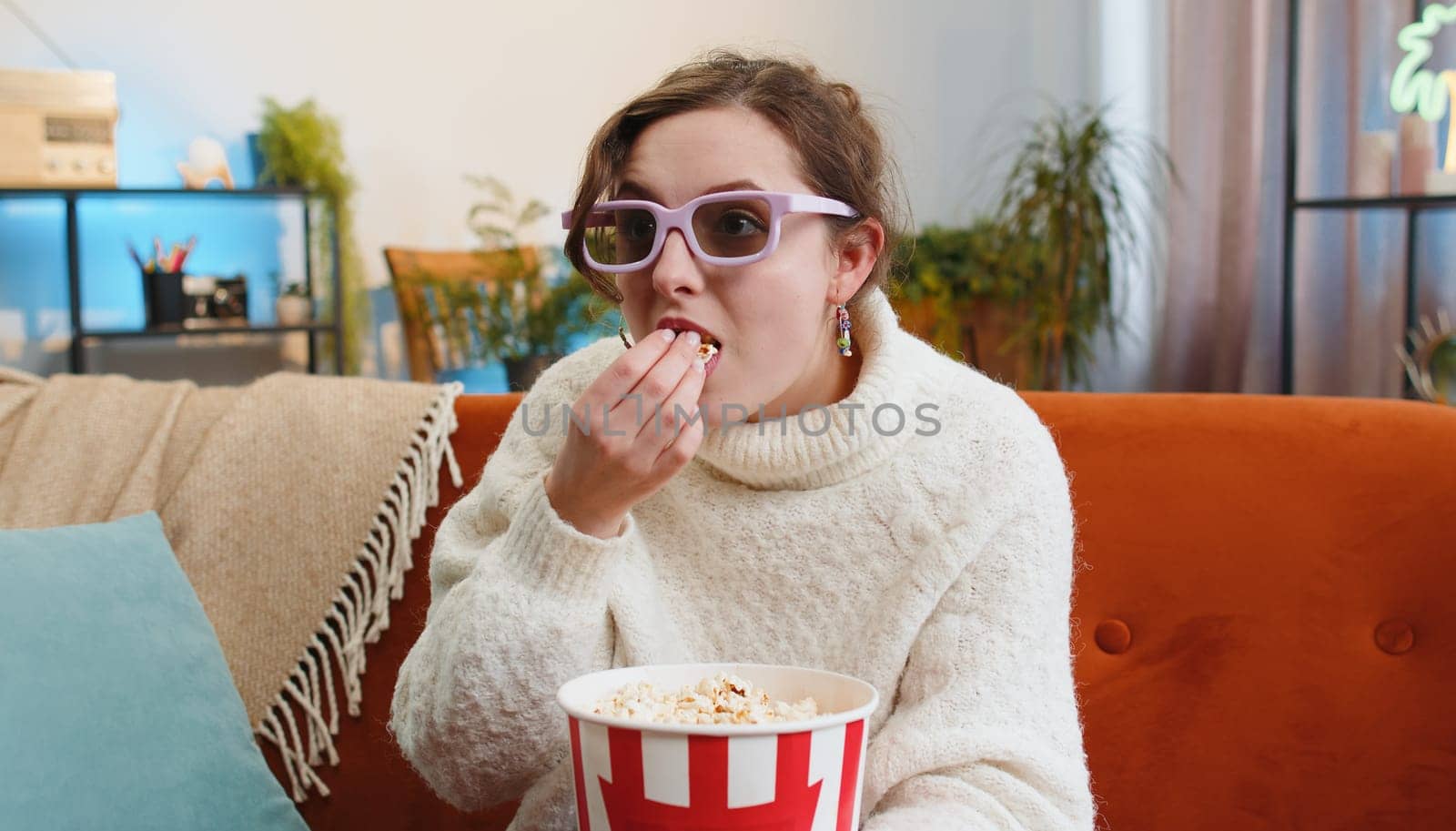 Excited young woman sitting on sofa eating popcorn and watching interesting tv serial, sport game, film, online social media movie content at home. Girl in 3D glasses enjoying domestic entertainment