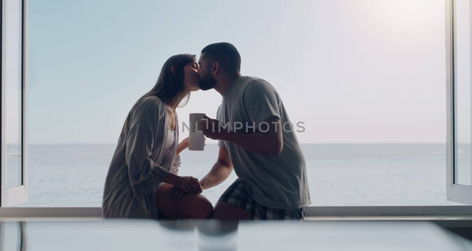 Couple, kiss and coffee in morning by window with love, romance and care on holiday by ocean with sunshine. Man, woman and trust with drink, espresso or matcha on vacation by sea with bond in summer.