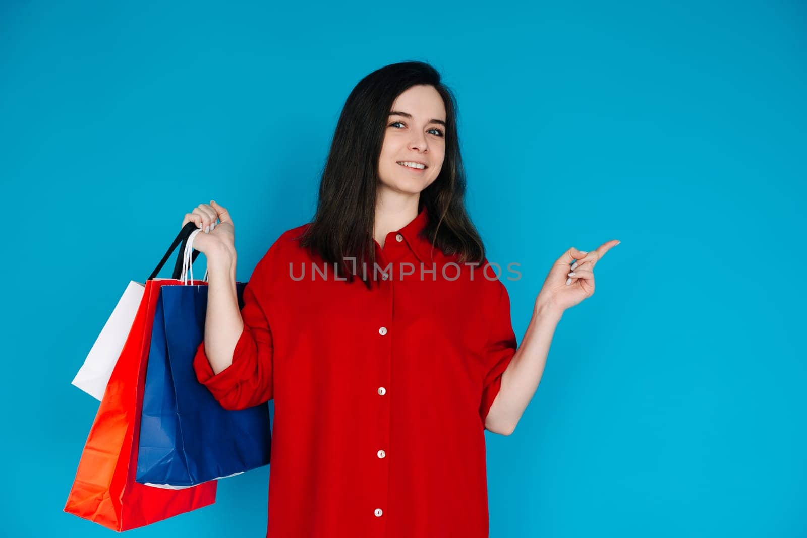 Cute Woman in Red Shirt with Shopping Bag - Pointing to Empty Space - Perfect for Promoting Sales and Discounts - Isolated on Blue Background. Smiling Woman with Shopping Bag