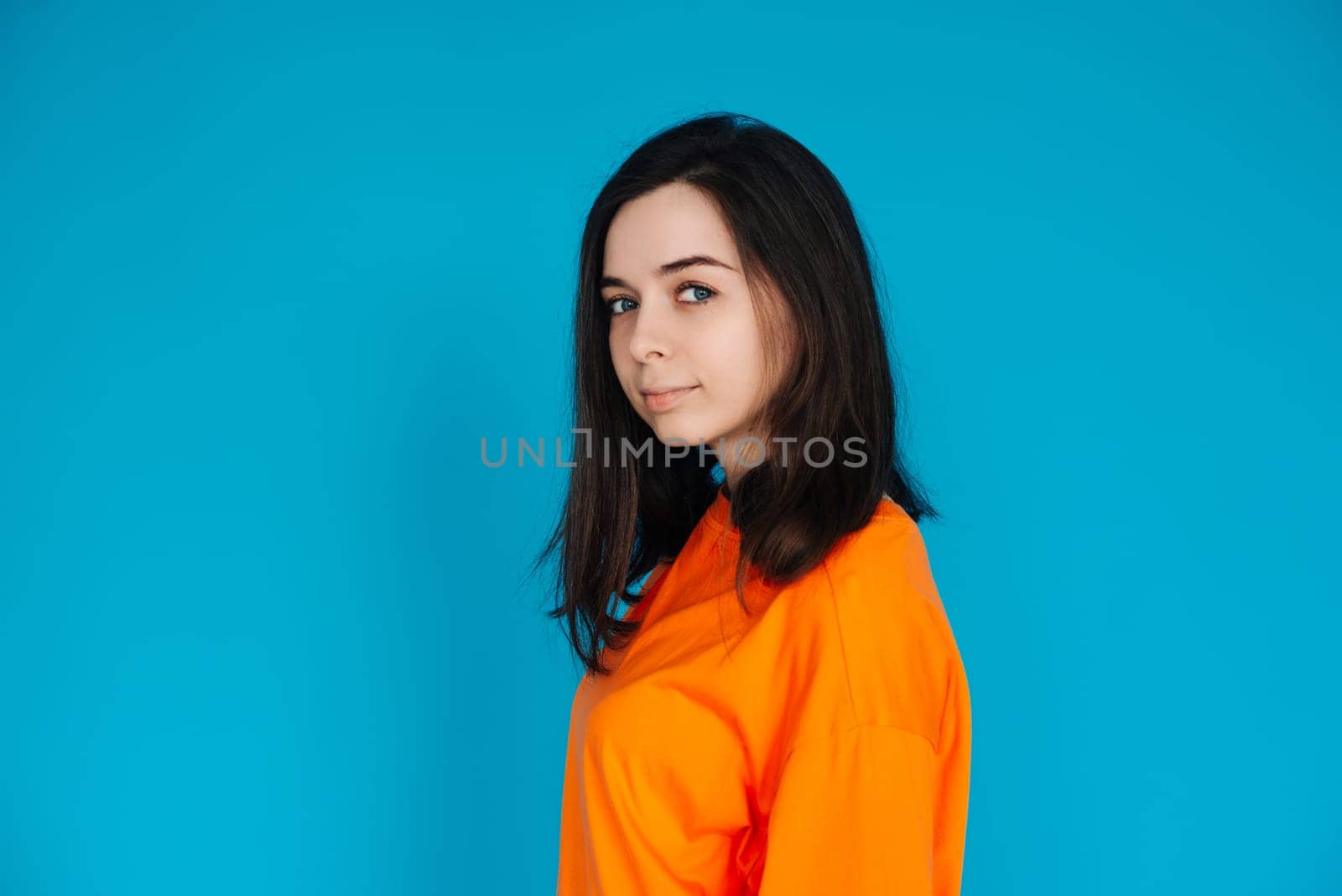 Stunning Young Woman in Trendy Orange Outfit Poses for Portrait, Isolated on Blue Background, Lovely Model with a Captivating Gaze and Gorgeous Appearance.