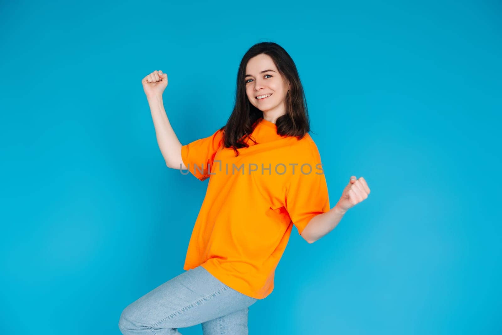 Excited Young Woman Celebrating Huge Sales Promotion, Raising Fists in Victory, Trendy Orange Outfit, Isolated on Blue Background, Portrait Photo.