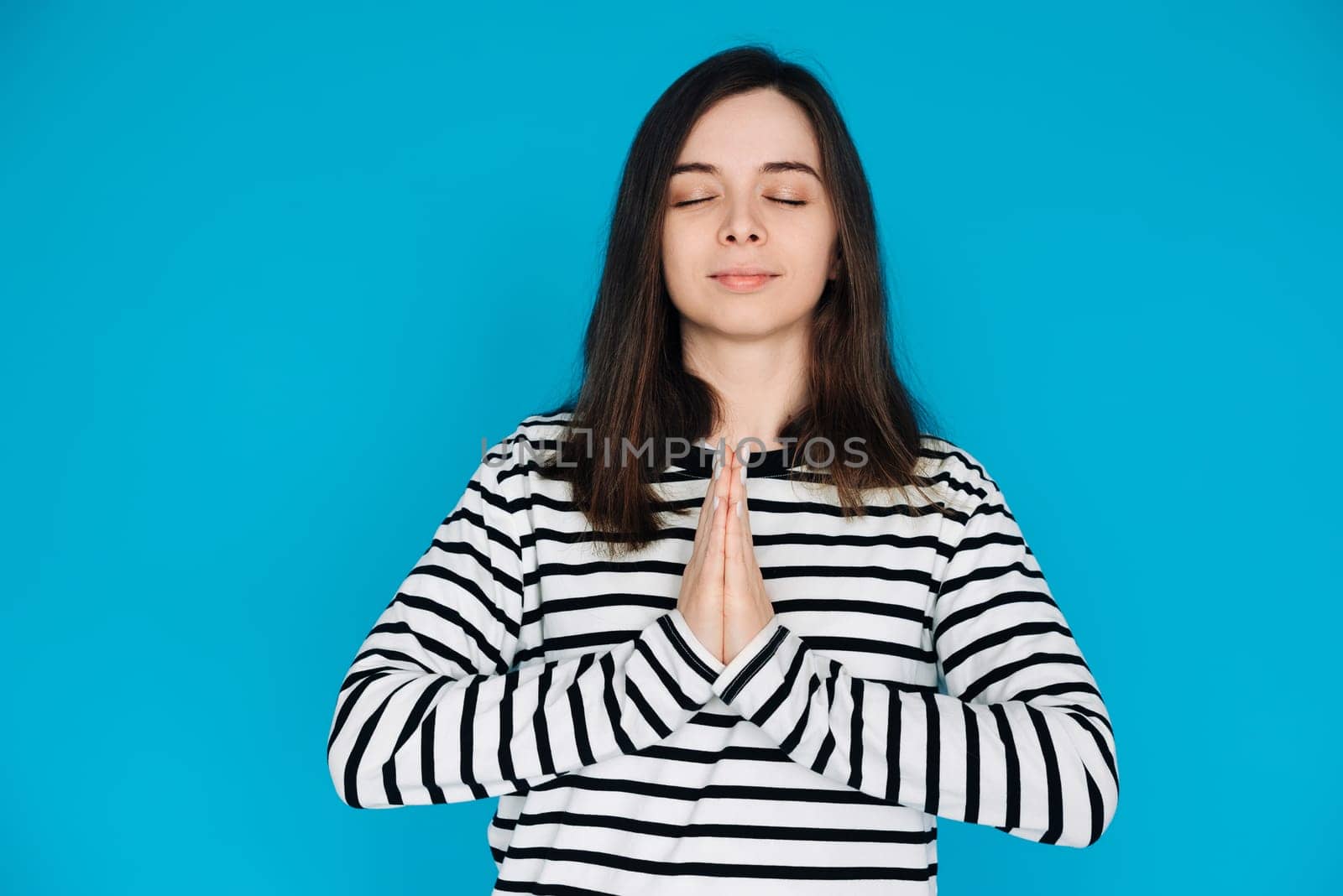 Serene Portrait of a Beautiful Young Woman Meditating with Closed Eyes and Palms, Inner Peace and Relaxation, Isolated on Blue Background.