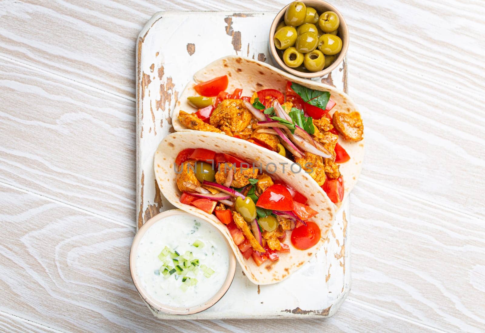 Traditional Greek Dish Gyros: Pita bread Wraps with vegetables, meat, herbs, olives on rustic wooden cutting board with Tzatziki sauce, olive oil top view, white wooden summer background.