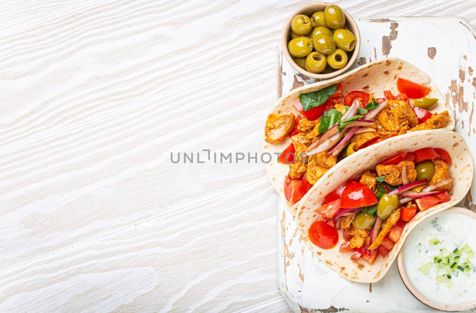 Traditional Greek Dish Gyros: Pita bread Wraps with vegetables, meat, herbs, olives on rustic wooden cutting board with Tzatziki sauce, olive oil top view on white wooden background, space for text.