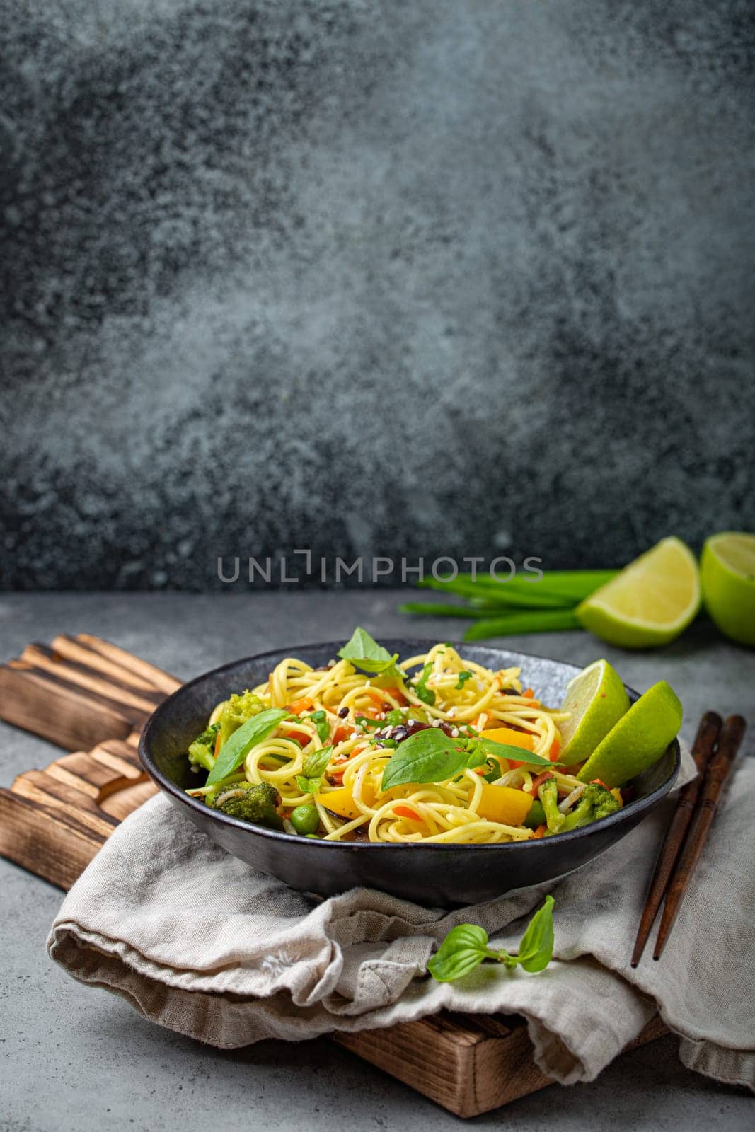 Asian vegetarian noodles with vegetables and lime in black rustic ceramic bowl, wooden chopsticks on cutting board angle view on stone background. Cooking noodles concept