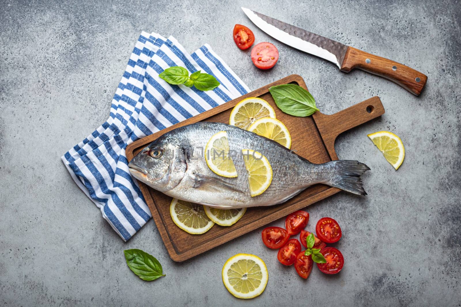 Raw uncooked fish dorado with ingredients lemon, fresh basil, cut cherry tomatoes on wooden cutting board with knife on rustic stone background top view, cooking healthy fish dorado concept