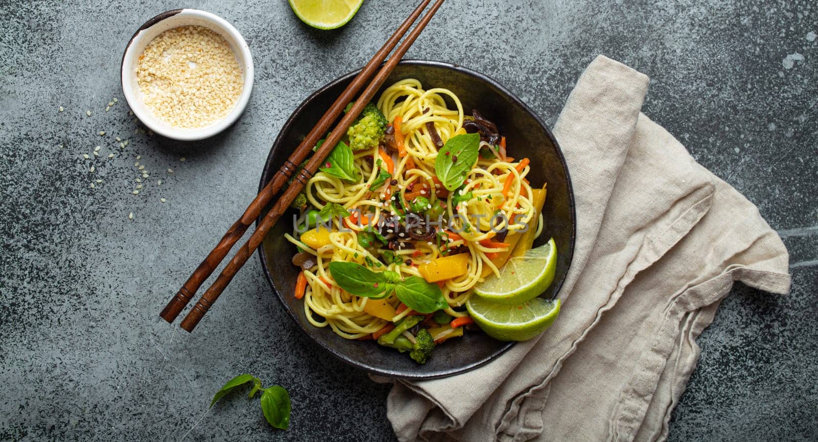 Asian vegetarian noodles with vegetables and lime in black rustic ceramic bowl, wooden chopsticks, cutting board with chopped green onion top view on stone background. Cooking noodles concept